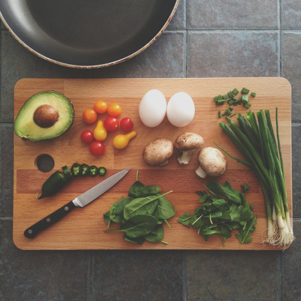 A wooden cutting board with a knife and prepped vegetables ready for cooking